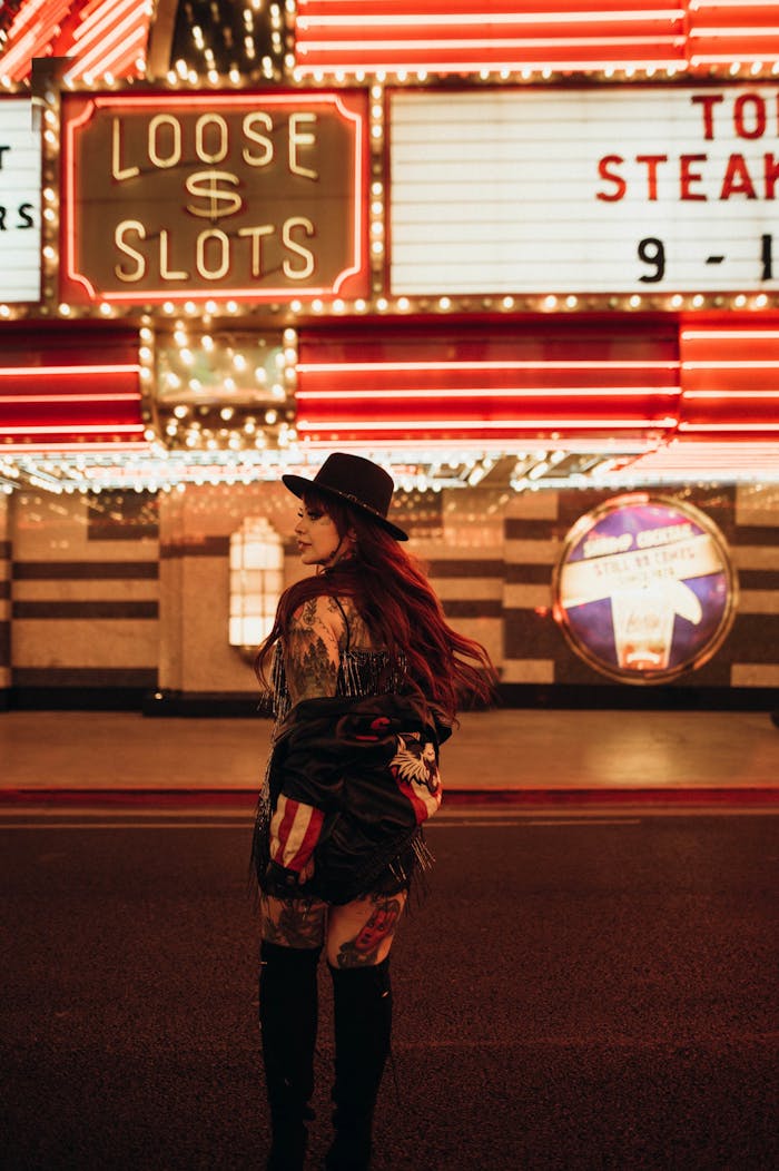 Tattooed woman in hat stands under neon lights at night in a city casino setting.